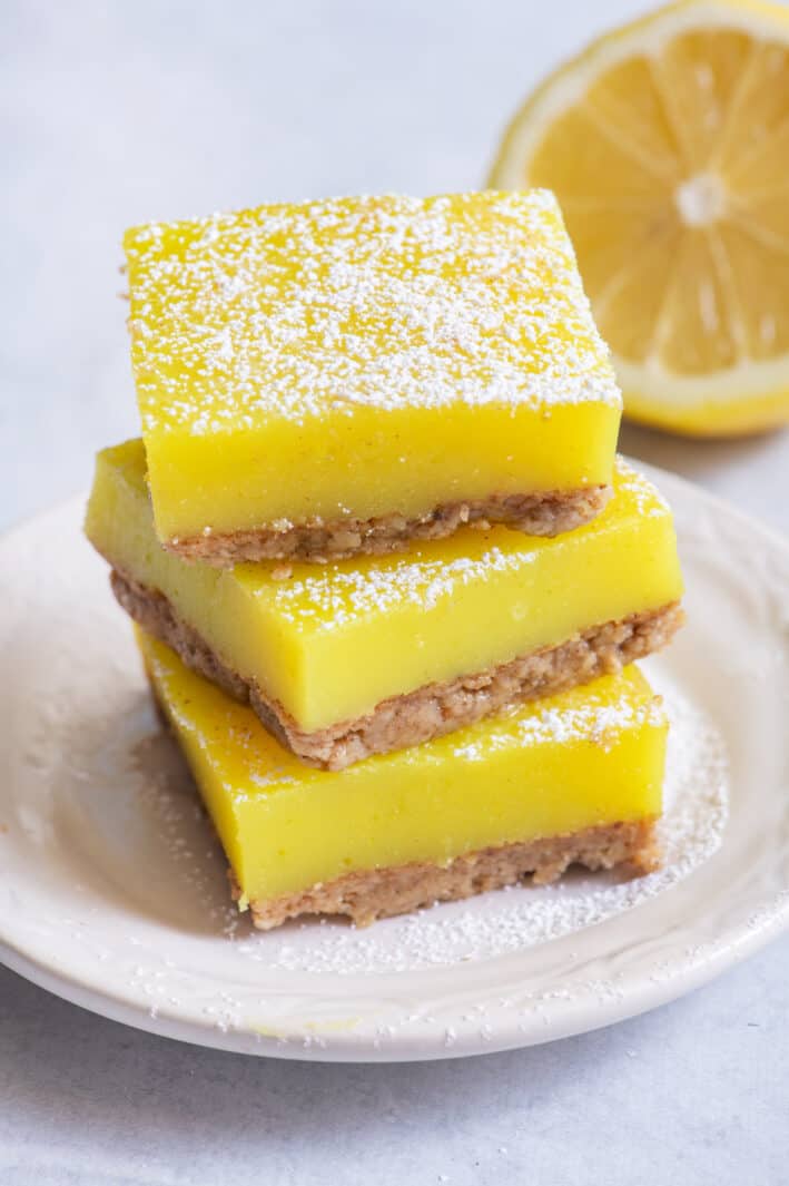 Stack of three vegan lemon bars on small white dish with half lemon in background