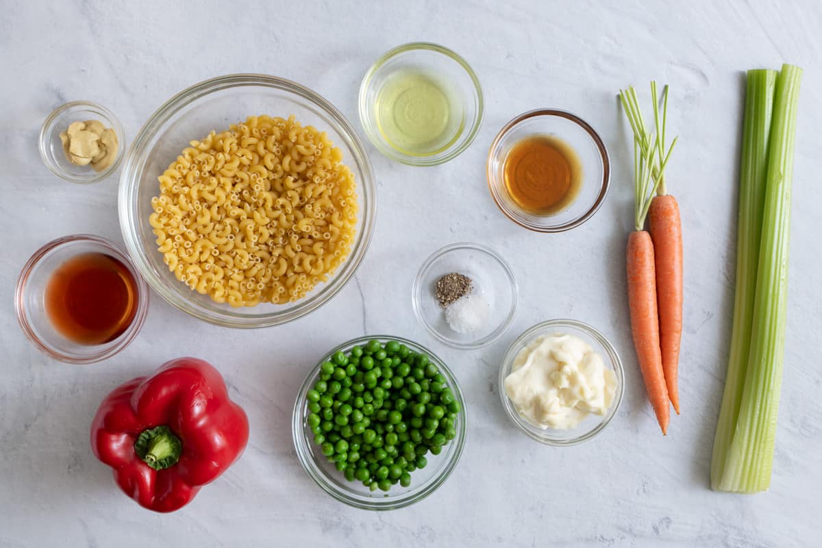 Ingredients for recipe before being prepped: dijon mustard, maple syrup, uncooked elbow pasta, whole red pepper, pickle juice, red wine vinegar, seasonings, frozen peas, vegan mayo, 2 whole carrots, and two celery stalks.