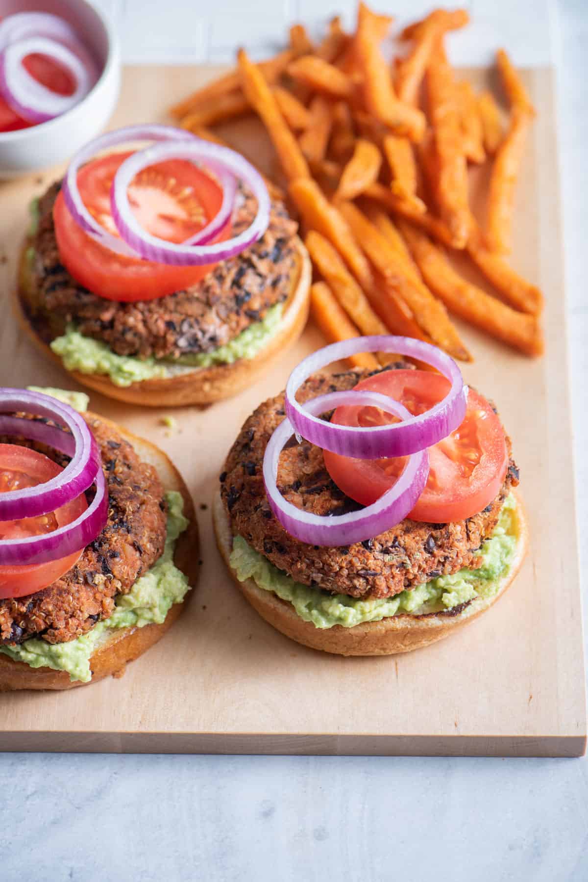 Three opened quinoa burgers topped with tomatoes and onions with sweet potato fries in background