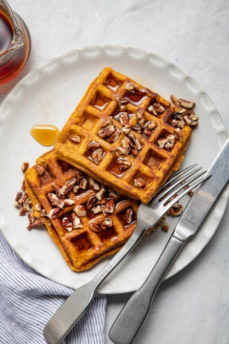 Sweet potato waffles stack on small white plate with fork and knife next to it.