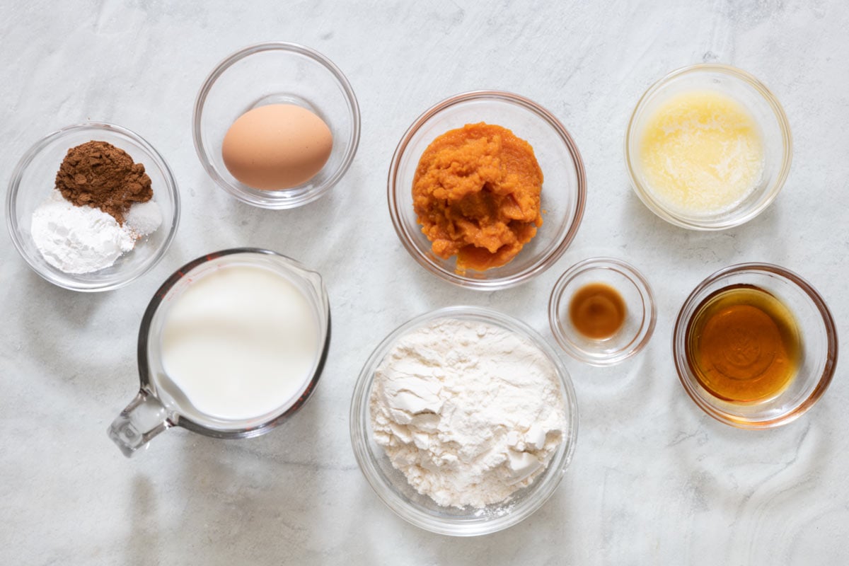 Ingredients before being prepped in individual bowls and cups: spices, a shelled egg, milk, pumpkin puree, flour, vanilla, melted butter, and maple syrup.