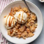 Apple crisp served with two scoops of vanilla ice cream and caramel sauce and spoon inside bowl