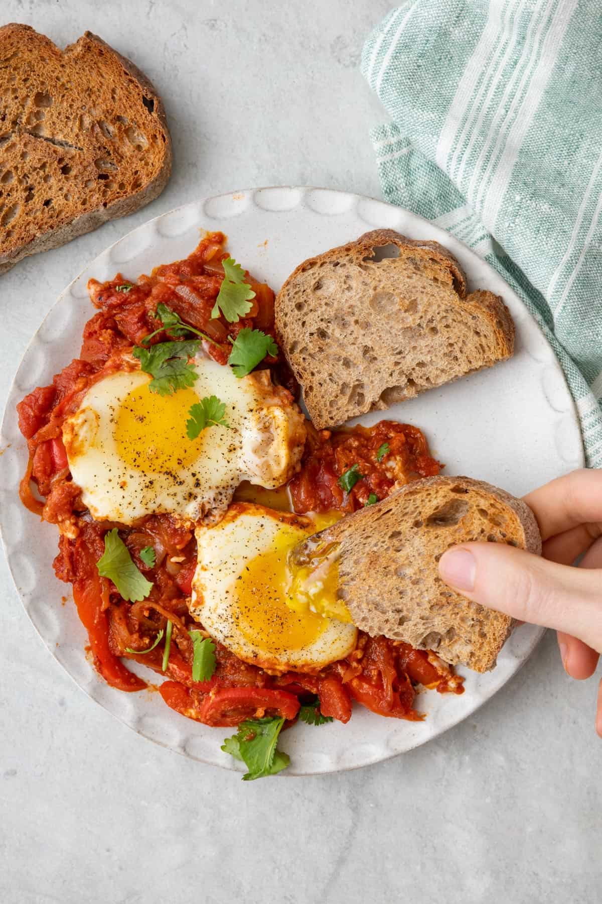 Two eggs served over shakshuka filling on a plate with a piece of toasted sourdough artisan bread dipping into the jammy egg yolk.
