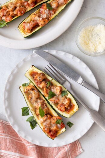 Final plating of the stuffed zucchini boats topped with fresh basil with fork and knife on plat and a side of grated parmesan to the side.