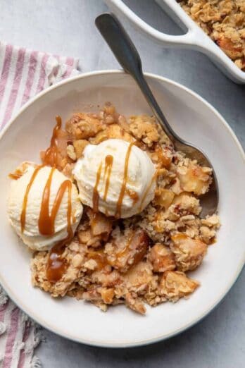 Apple crisp served with two scoops of vanilla ice cream and caramel sauce and spoon inside bowl