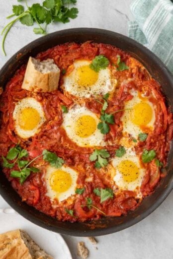 Shakshuka in pan garnished with cilantro and sourdough nearby with a piece in on top and showing cooked eggs in rich sauce.