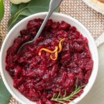 Bowl of cranberry orange sauce garnished with orange zest and fresh sprig of thyme with a spoon dipped in.