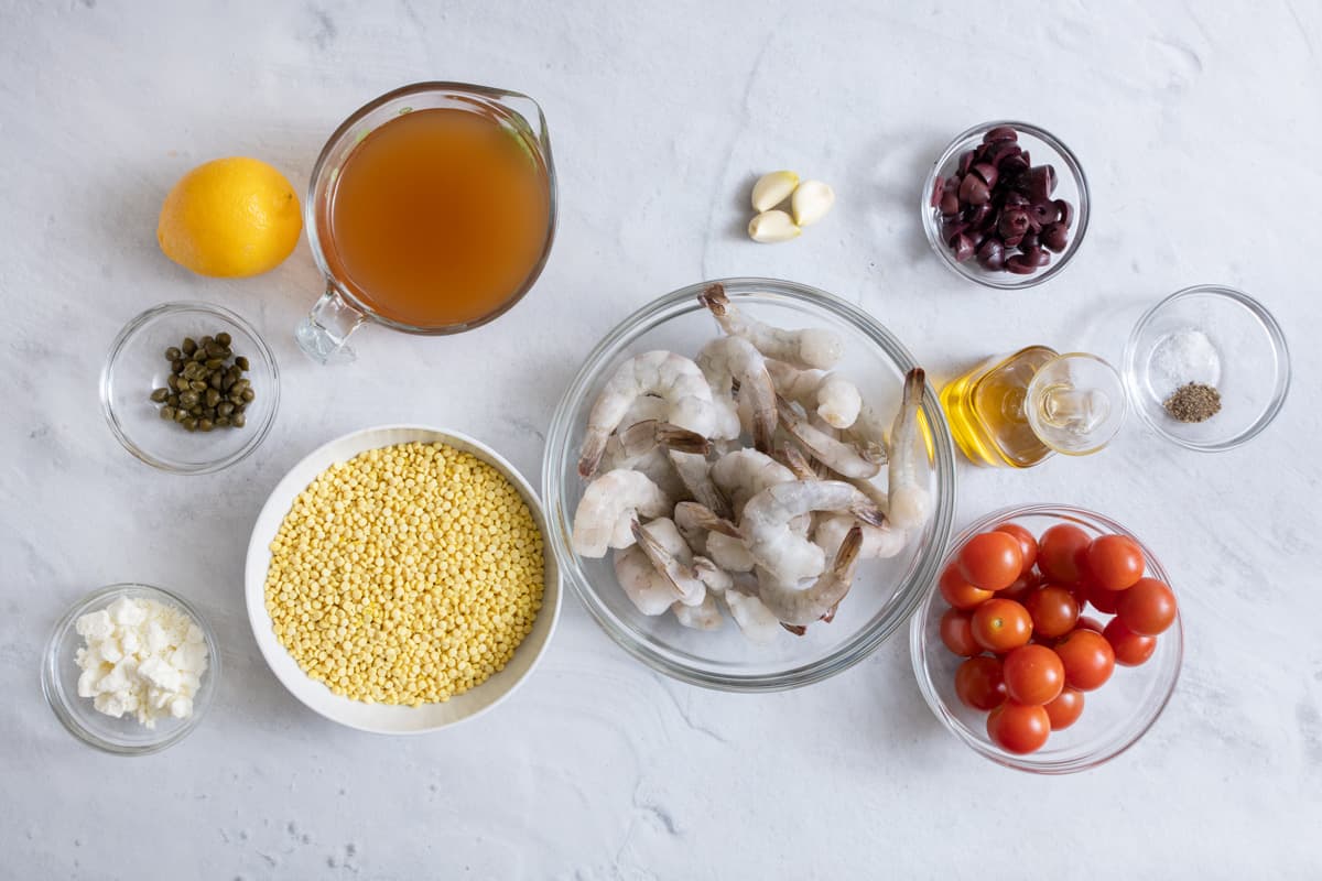 Ingredients for recipe before prepped: lemon, capers, feta, broth, uncooked couscous, raw shrimp, oil, spics, garlic cloves, and cherry tomatoes.