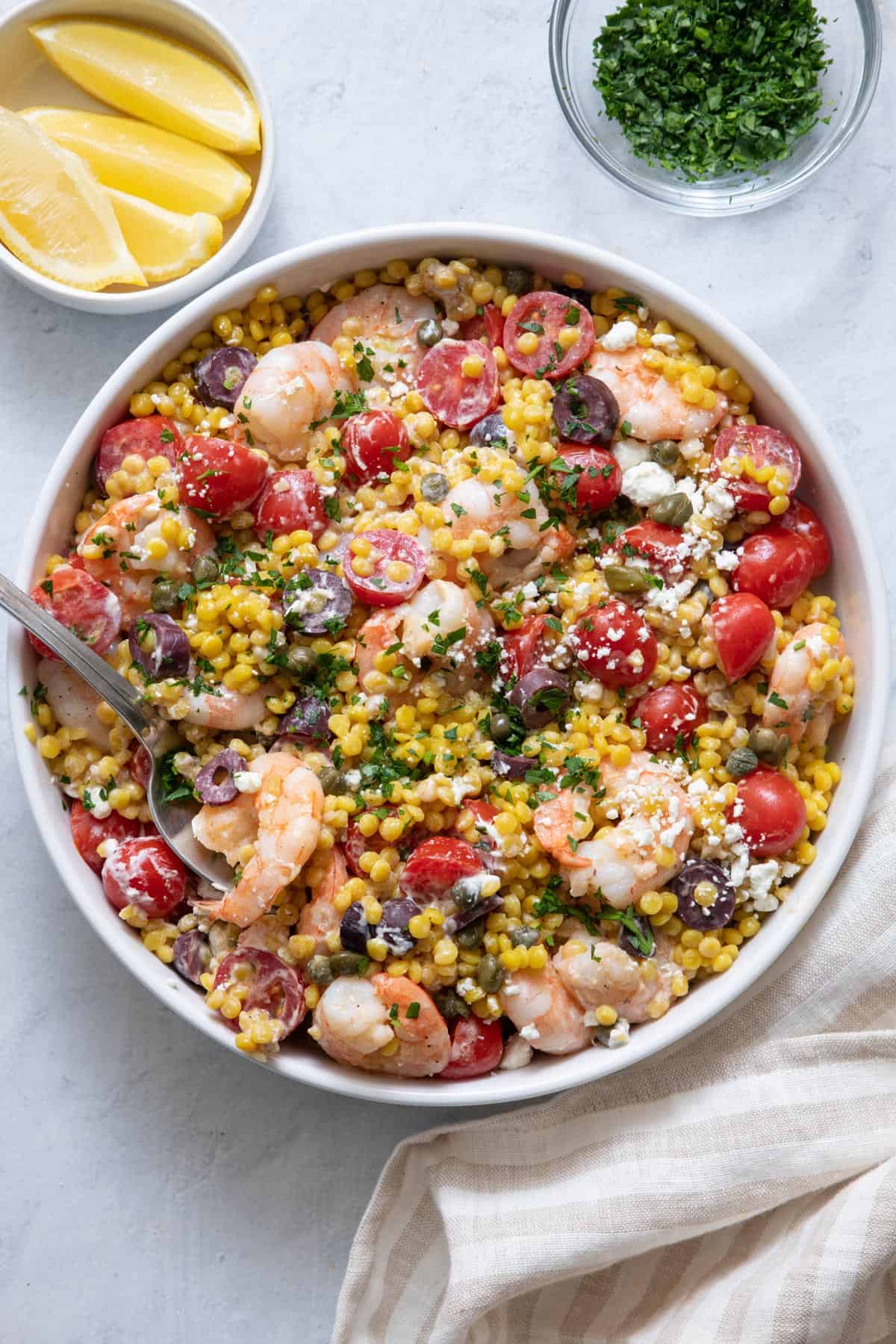 Close up shot of the Mediterranean Couscous in large white serving bowl with chopped parsley and lemon wedges off to side.