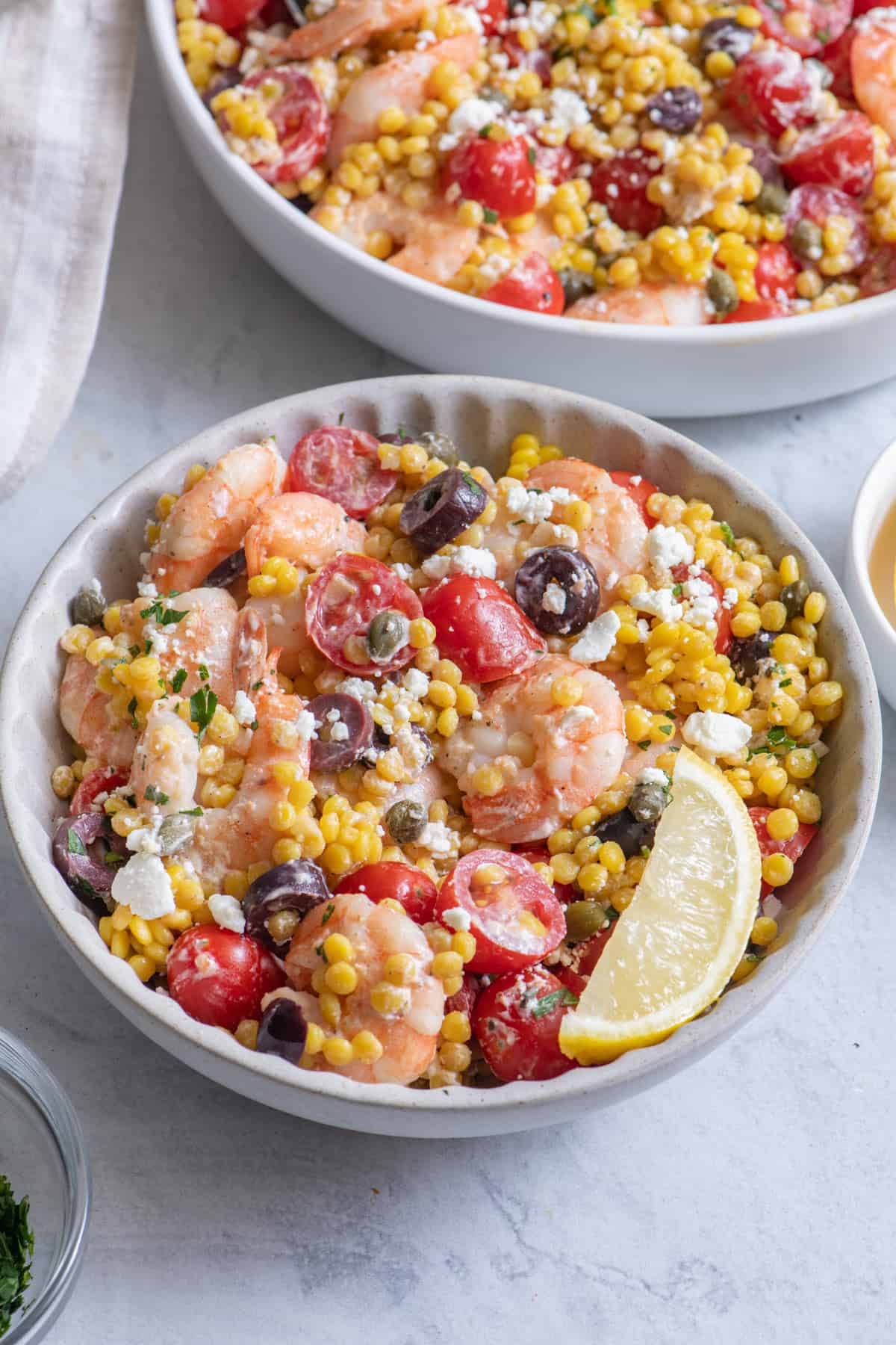 Close up serving of couscous shrimp salad recipe in white bowl with large serving dish in back.