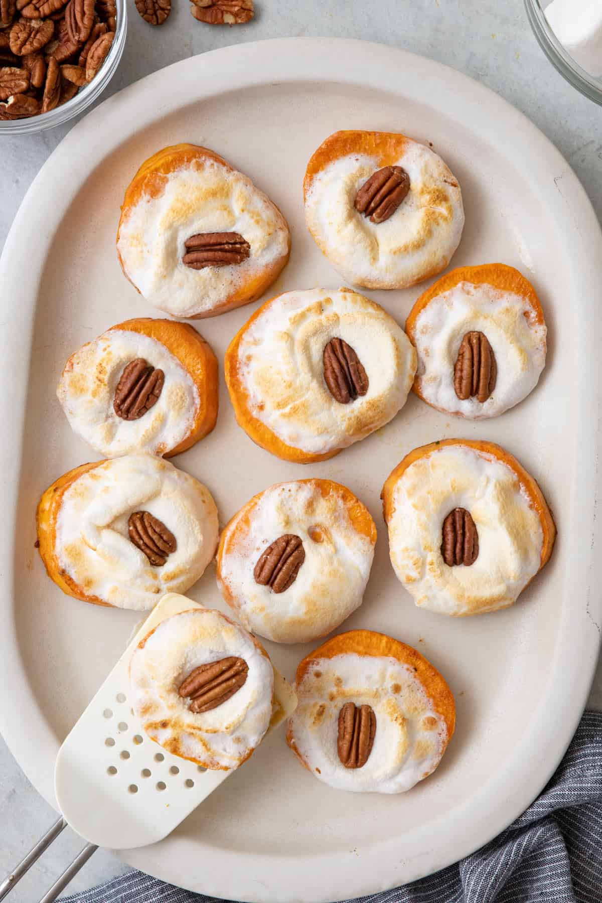 Sweet potato bites on a oval white dish with melted marshmallows and a pecan half on each with a spatula lifting one up.