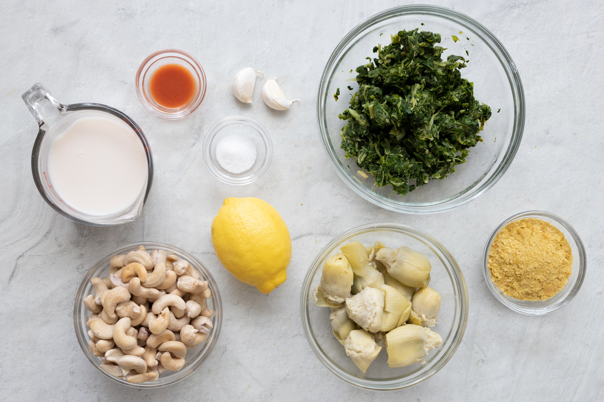 Ingredients for recipe in individual bowls: almond milk, whole cashews, hot sauce, salt, whole lemon, 2 garlic cloves, spinach, artichoke hearts, and nutritional yeast.