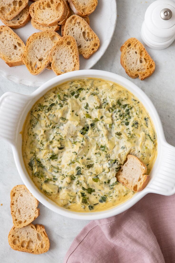 Overhead image of spinach artichoke dip after baked being served with a plate of toasted crostinis for dipping with one inside dip.