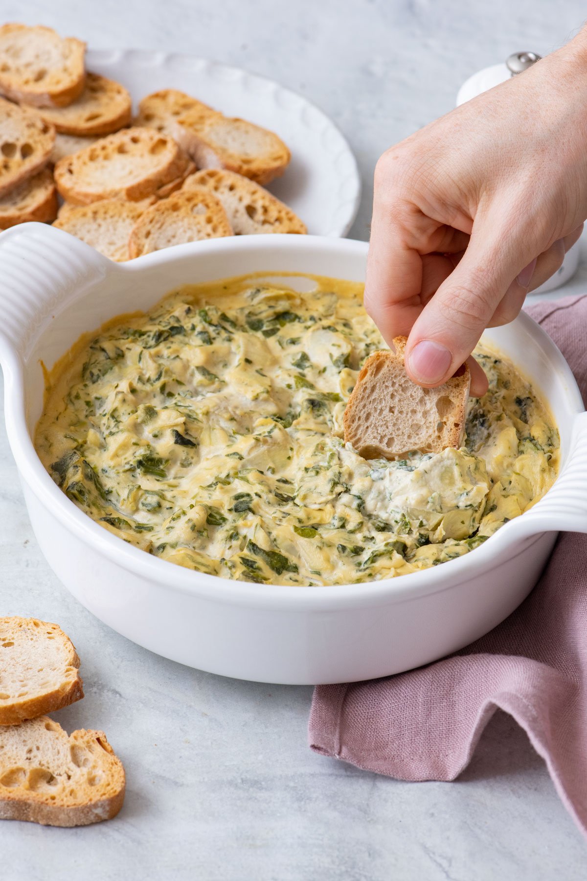 Small toast dipping into warm vegan spinach artichoke dip in round white casserole dish with more toasts nearby.