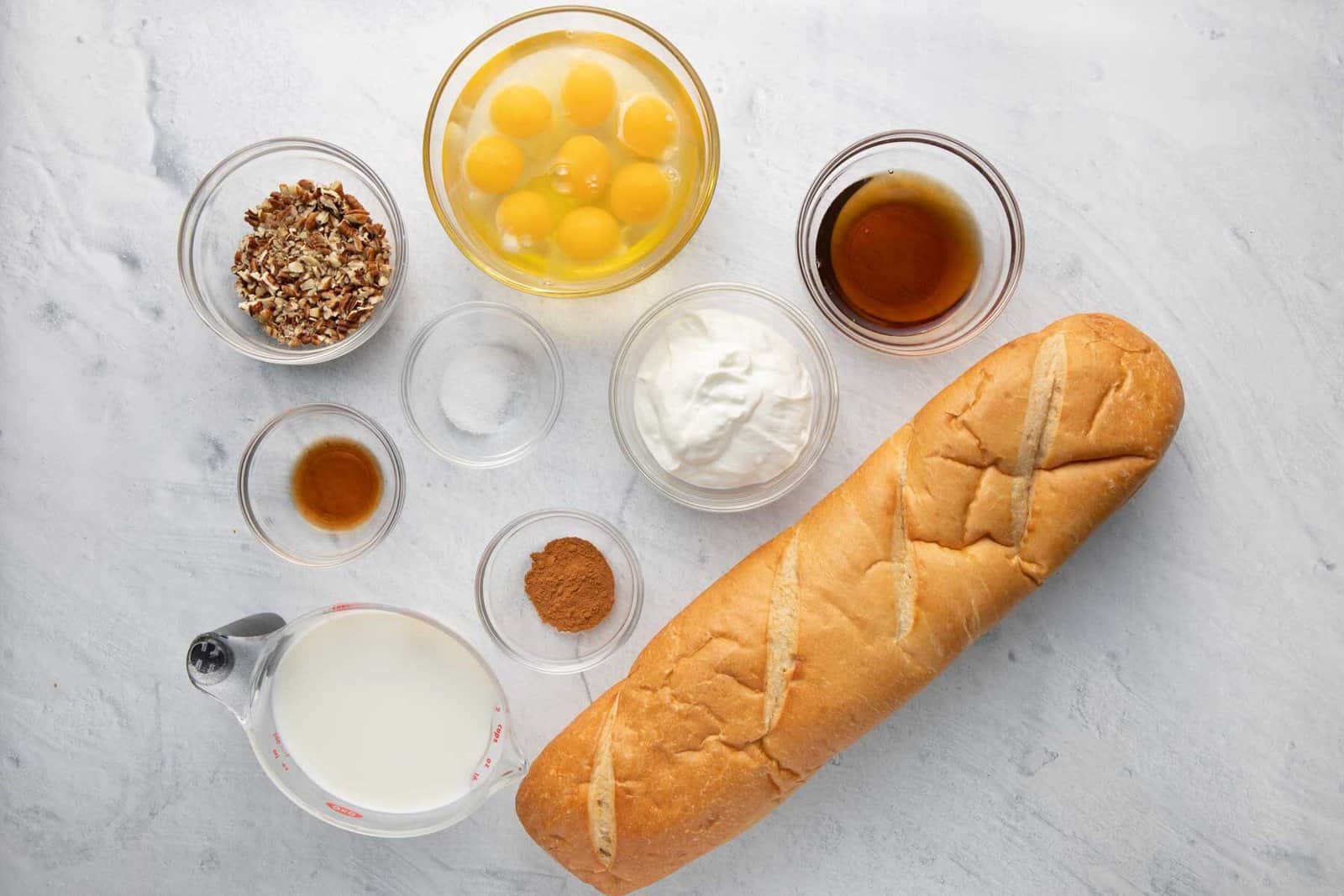 Ingredients for recipe in individual bowls: chopped pecans, vanilla, milk, salt, cracked eggs, cinnamin, yogurt, maple syrup, and a loaf of bread.