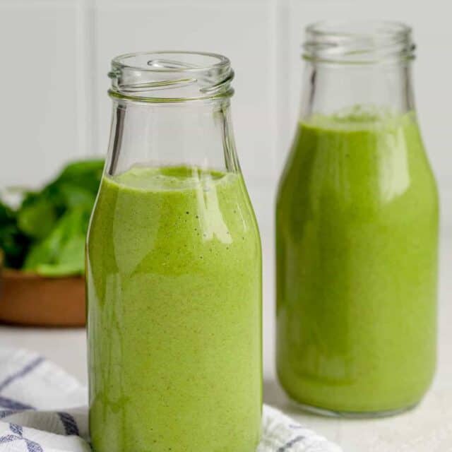 Two small milk jugs of green smoothies with bowl of spinach in background