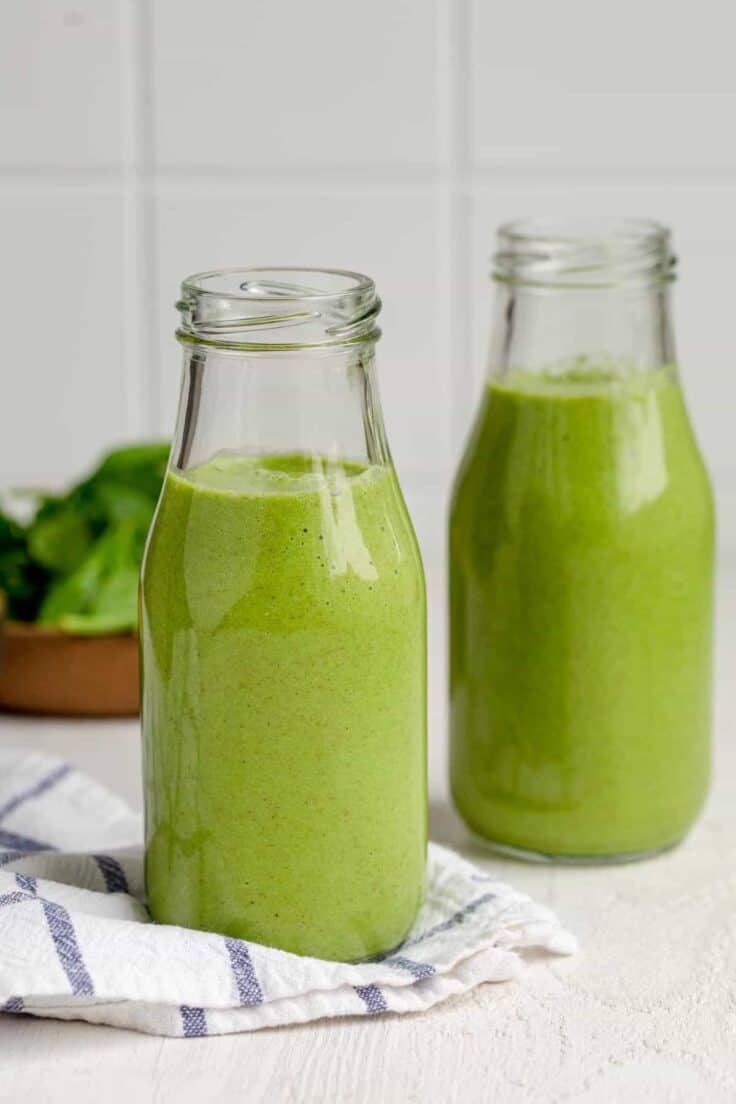 Two small milk jugs of green smoothies with bowl of spinach in background