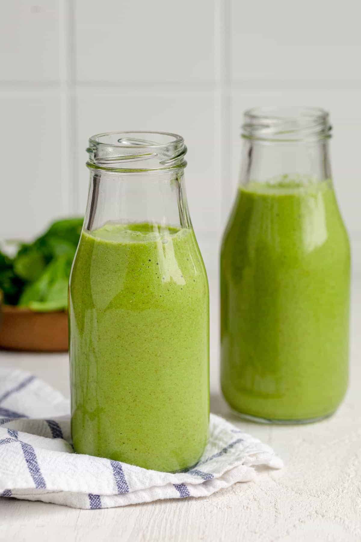 Two small milk jugs of green smoothies with bowl of spinach in background