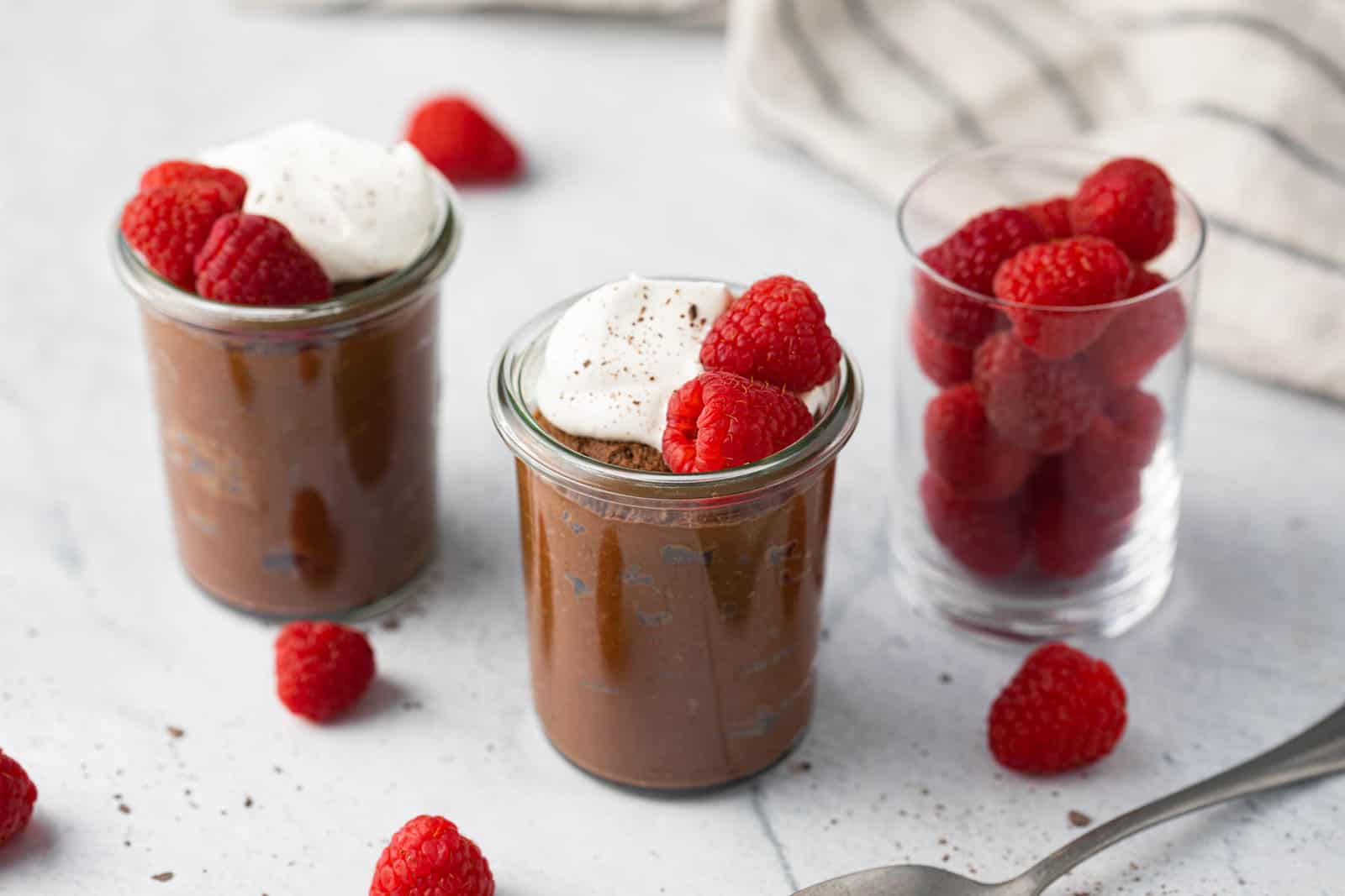 Two small jars of avocado chocolate mousse topped with whipped cream and fresh raspberries with a small jar filled with fresh raspberries.