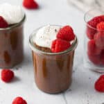 2 small jars of chocolate avocado mouse with whipped cream and raspberries with a closer shot on the center jar, and a small glass jar on the other side filled with fresh raspberries. A spoon and more fresh berries scattered around.