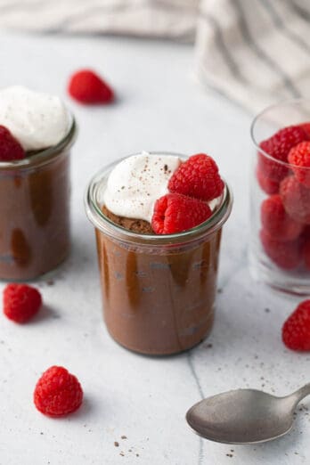 2 small jars of chocolate avocado mouse with whipped cream and raspberries with a closer shot on the center jar, and a small glass jar on the other side filled with fresh raspberries. A spoon and more fresh berries scattered around.