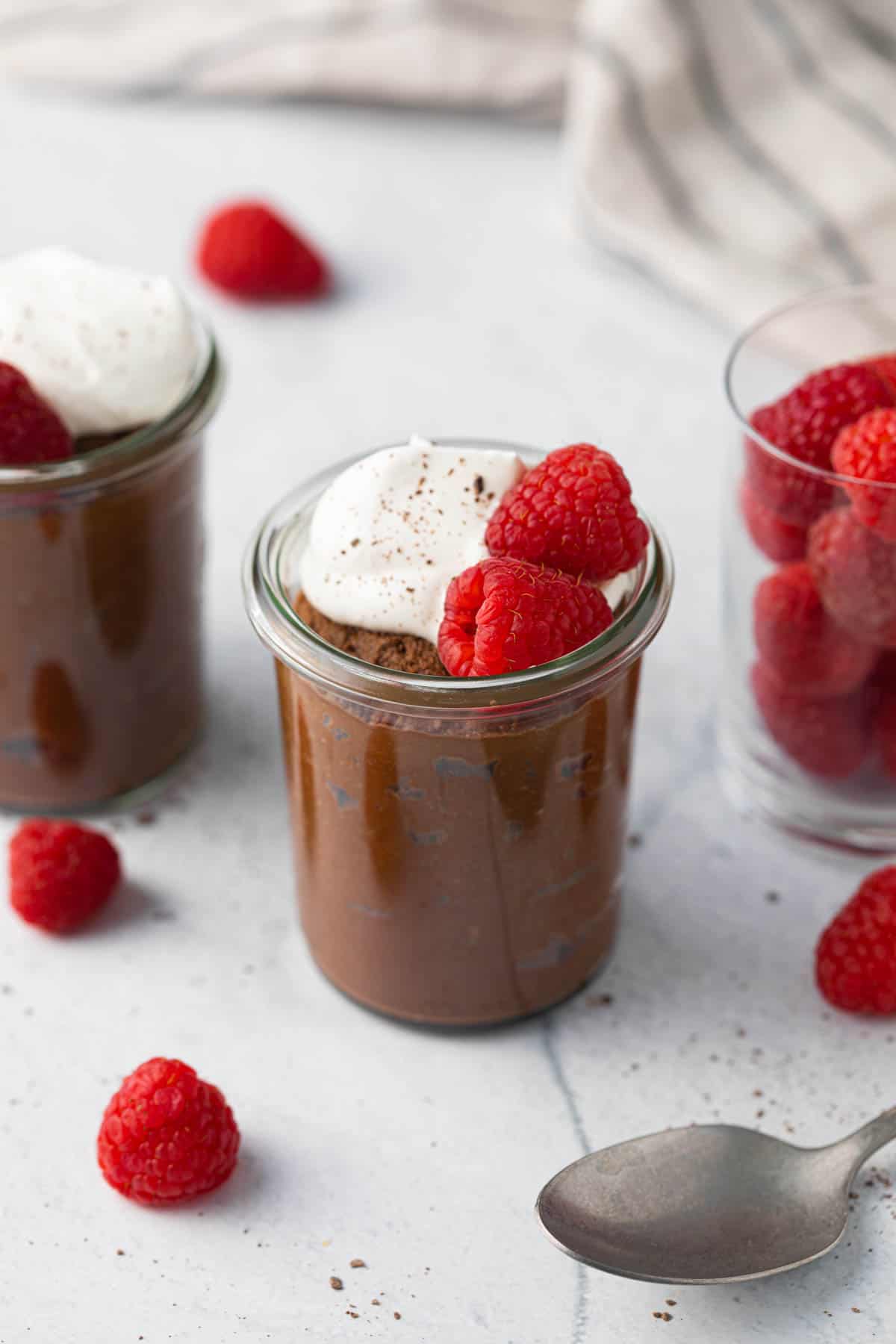 2 small jars of chocolate avocado mouse with whipped cream and raspberries with a closer shot on the center jar, and a small glass jar on the other side filled with fresh raspberries. A spoon and more fresh berries scattered around.
