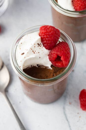 Close view of avocado chocolate mousse served in a glass jar topped with whipped cream and fresh raspberries with a bite taken out and another jar of recipe off to the side.