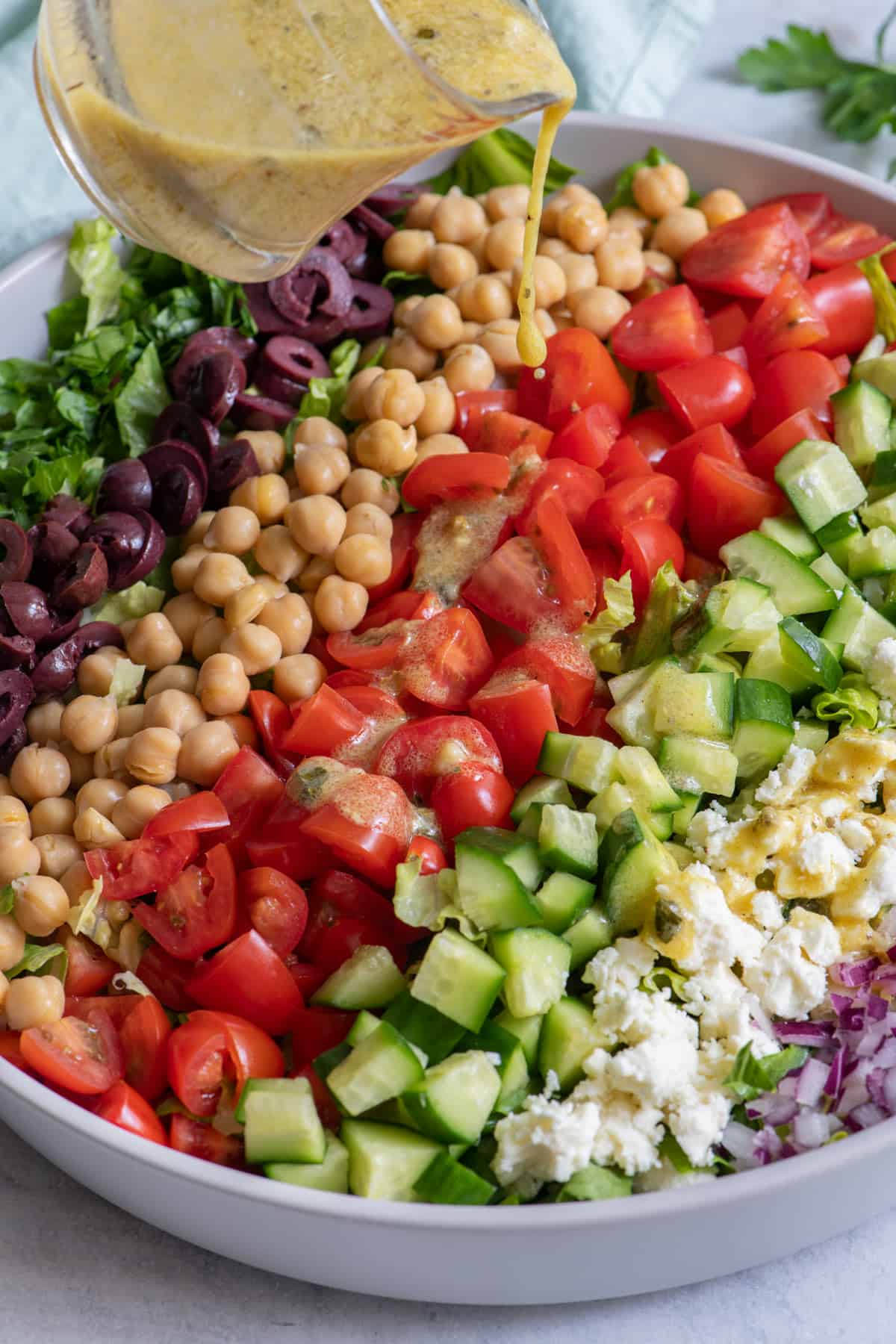 Dressing poured over a big bowl of salad with lettuce, olives, chickpeas, chopped tomatoes, diced cucumbers, feta, and diced red onions.