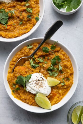 Two bowls of red lentil curry over rice with cilantro, yogurt and lime wedges