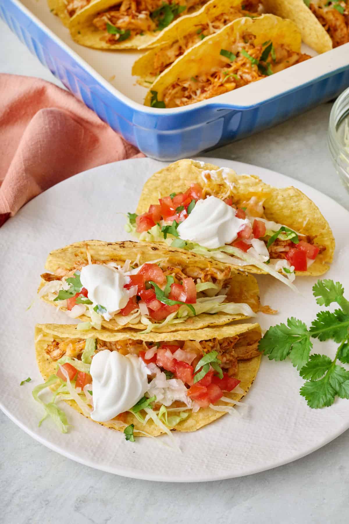 3 prepared baked chicken tacos on a small plate garnished with sour cream, tomatoes, and fresh cilantro with baking dish nearby.