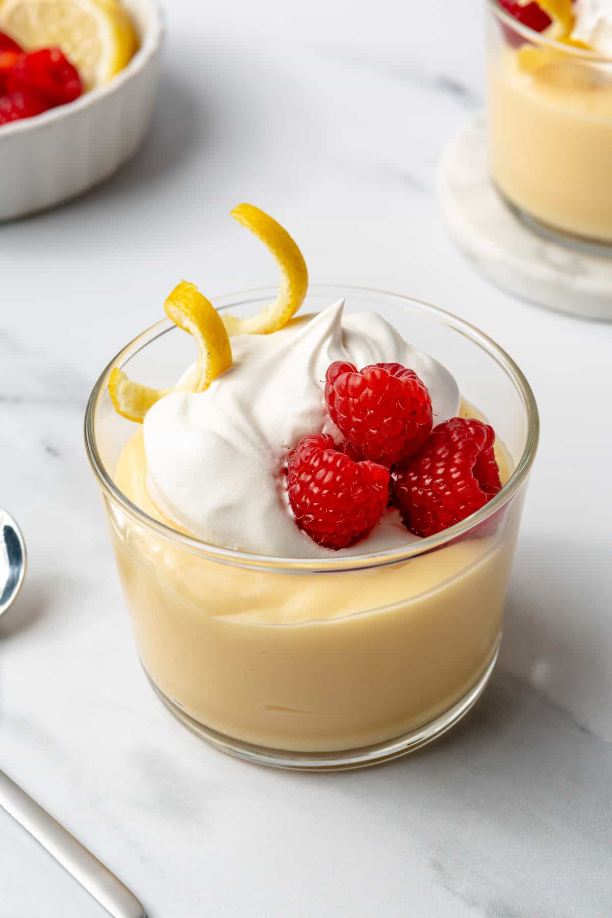 Small dish of lemon pudding topped with whipped cream, fresh raspberries, and lemon zest.