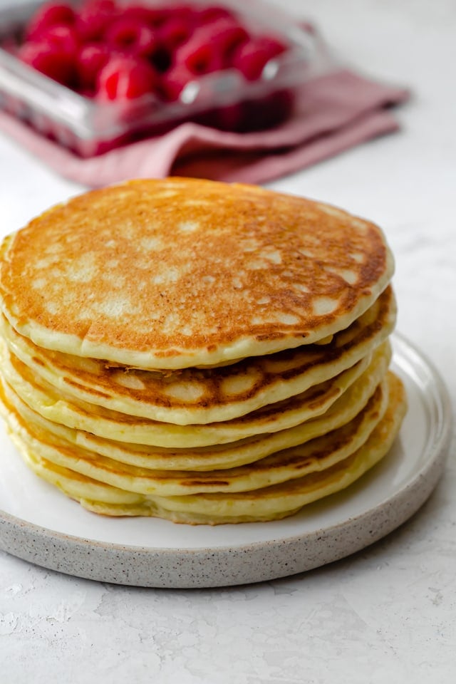 Stack of lemon ricotta pancakes on a white plate with raspberries in the background