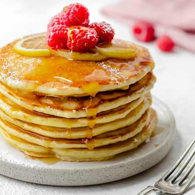 Stack of lemon ricotta pancakes with maple syrup drizzling down the sides and topped with raspberries