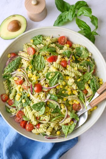Avocado pasta salad in a serving bowl with serving set dipped inside, halved avocado nearby.