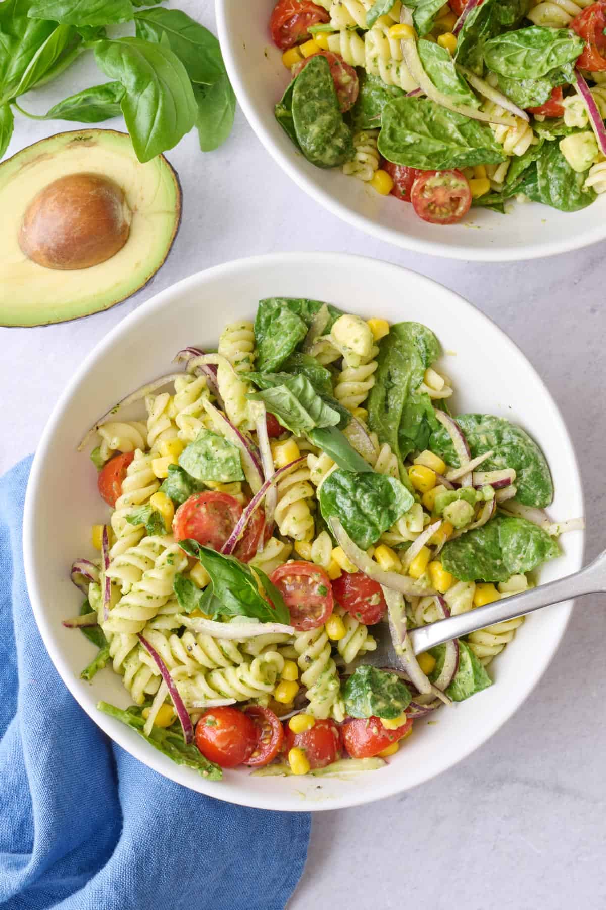 2 servings of pasta salad in shallow bowls garnished with fresh basil, halved avocado nearby.