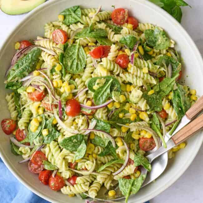 Avocado pasta salad in a serving bowl with serving set dipped inside, halved avocado nearby.