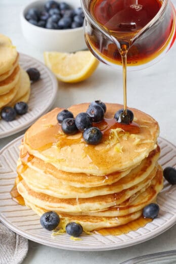 Syrup being poured over a stack lemon ricotta pancakes with another plate nearby. Fresh blueberries and lemon zest on top.