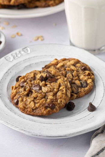 2 oatmeal raisin chocolate chip cookies on a small plate with a glass of milk nearby.