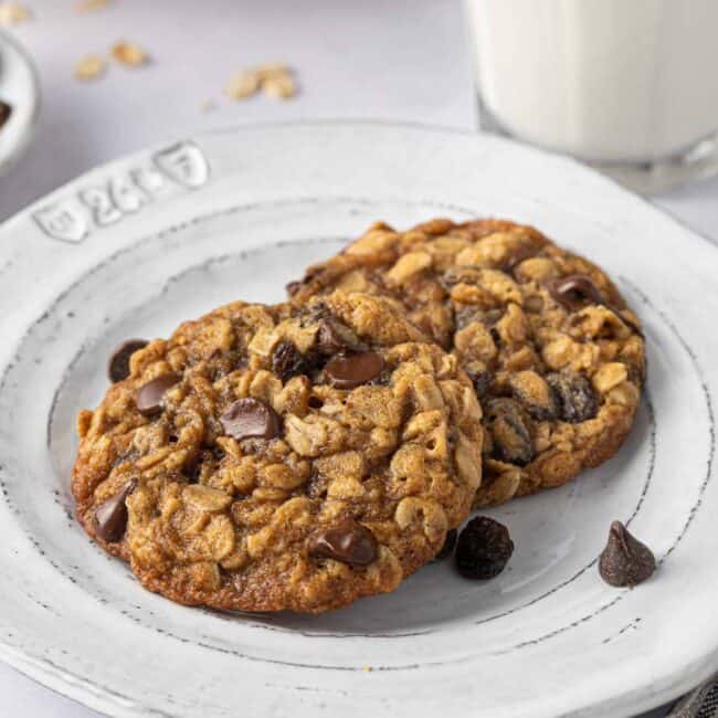 2 oatmeal raisin chocolate chip cookies on a small plate with a glass of milk nearby.