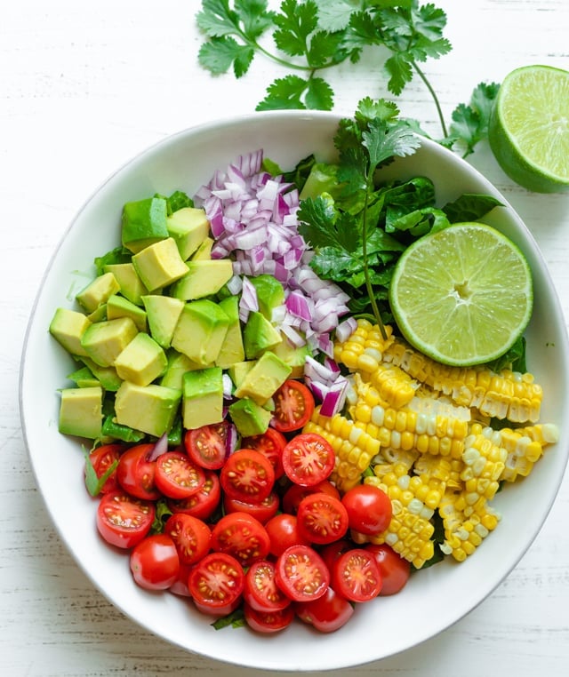 Ingredients to make the recipe: lettuce, corn, tomatoes, avocado, cilantro, red onions and lime juice pictured in a large bowl