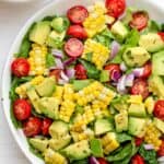 Corn tomato avocado salad in a large white bowl with squeezed limes on the side
