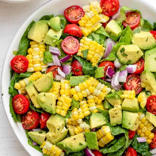 Corn tomato avocado salad in a large white bowl with squeezed limes on the side