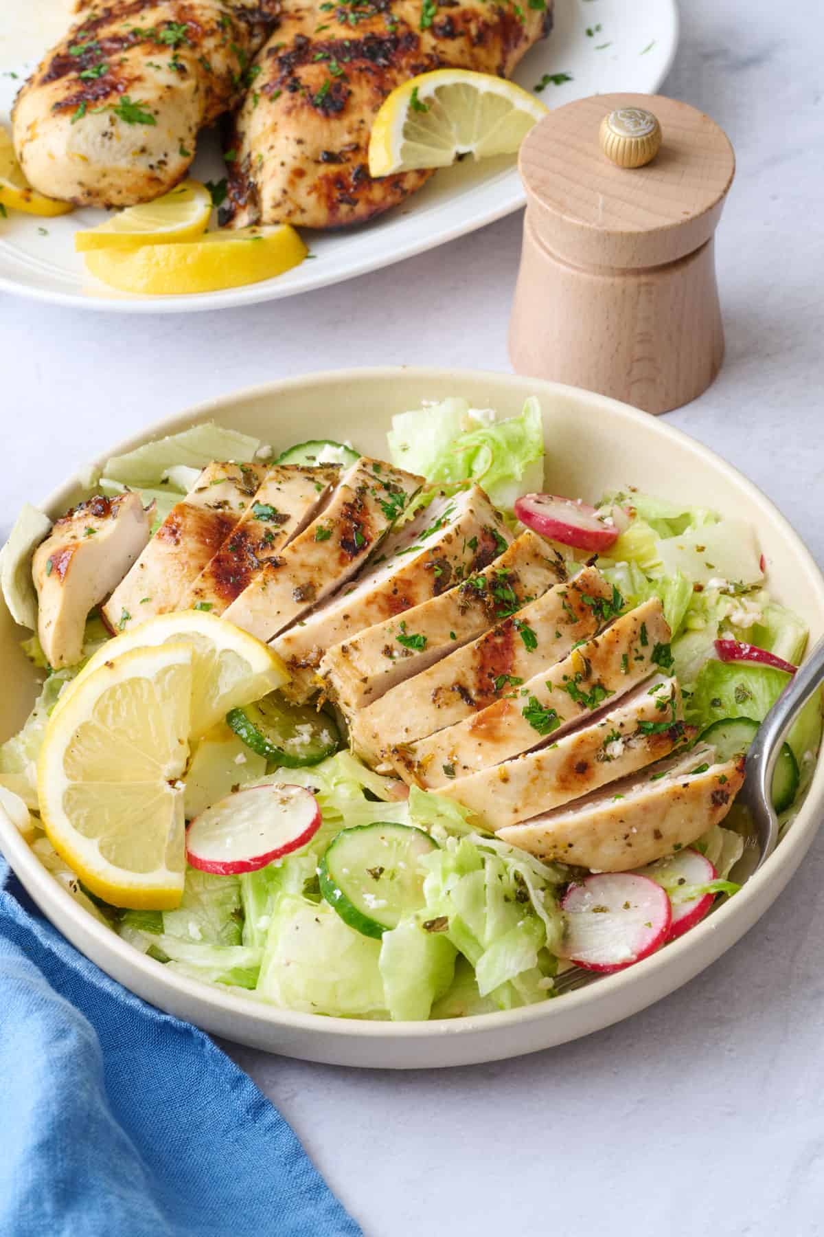 Sliced grilled chicken on a plate with the iceberg lettuce salad below and lemon wedges with platter nearby.