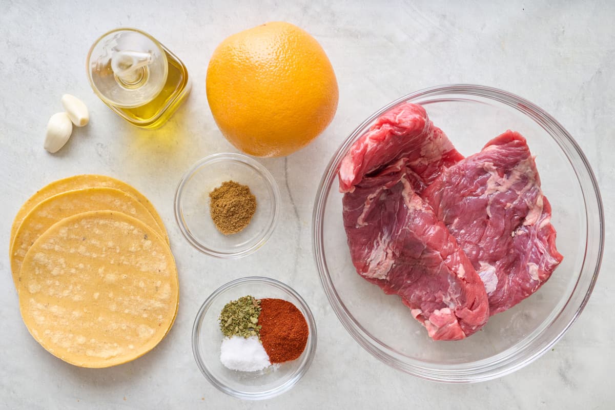 Ingredients for recipe before prepping: corn tortillas, garlic, oil, orange, spices, and steak.