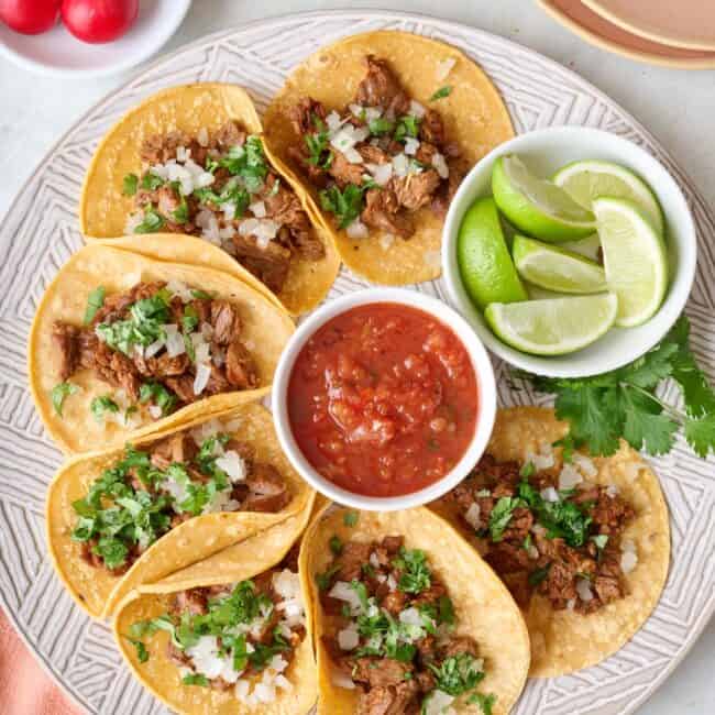 Homemade Mexican Street Tacos on a round platter with a small dish of salsa, garnished with fresh chopped cilantro and onions.