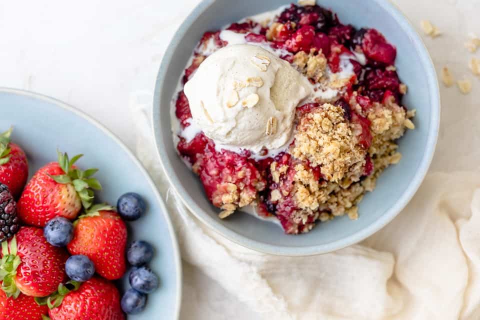 Crisp in a bowl topped with vanilla icecream, with a bowl of berries nearby.