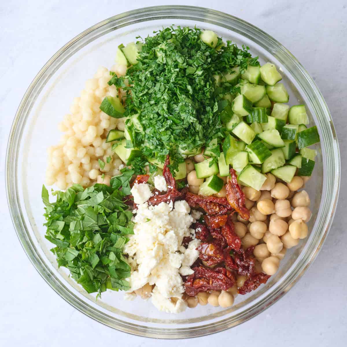 Cooled couscous added to the large bowl of dressing with cucumbers, chickpeas, sundried tomatoes, feta and herbs on top.