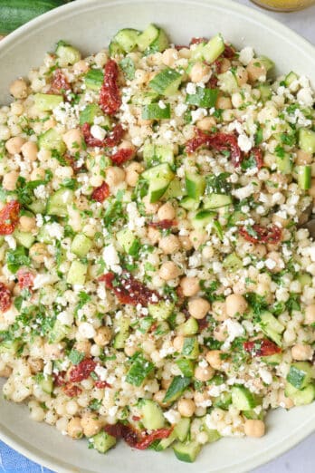 Couscous salad in a wide shallow bowl garnished with extra feta, serving spoon dipped inside with small jar of extra dressing nearby.