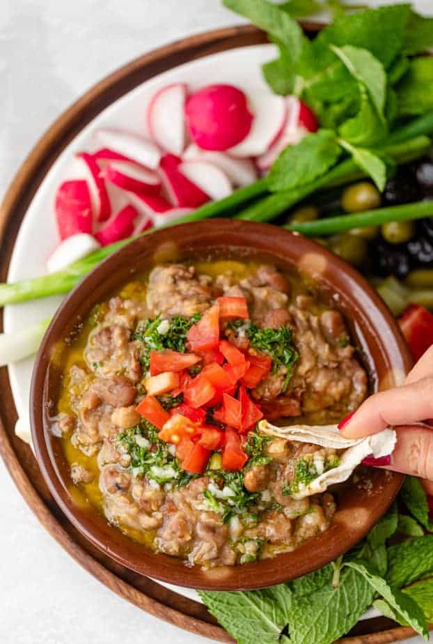 Pita dipping into ful medames bowl surrounded by vegetables