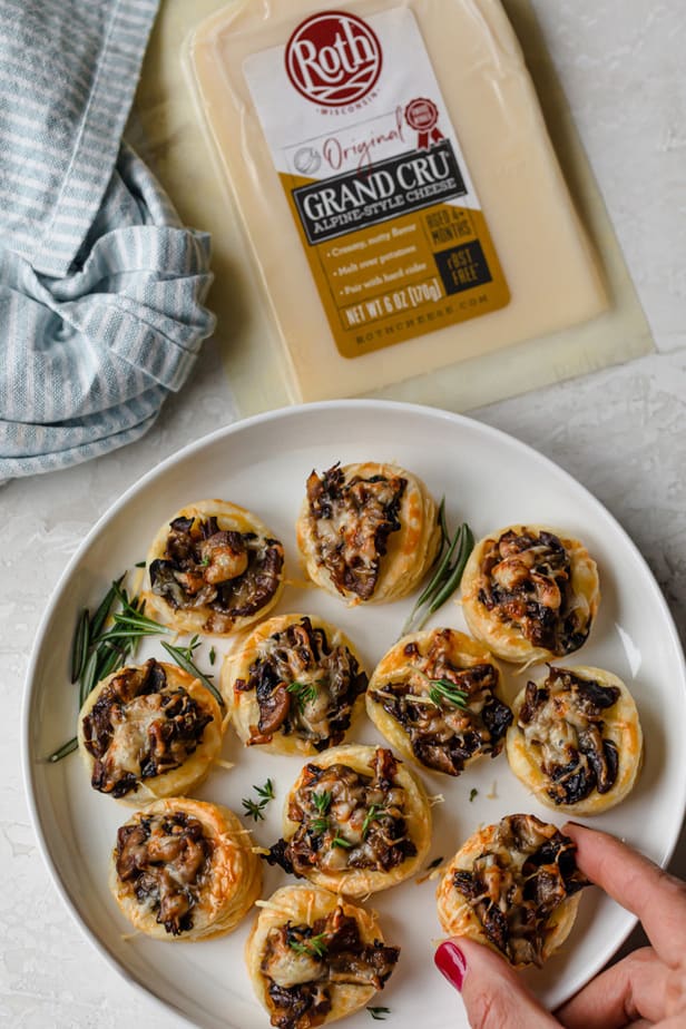 Mushroom tartlets in a large plate with hand grabbing one and Roth Cheese in the background